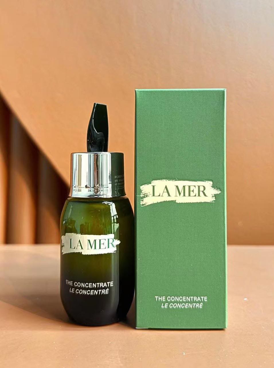 La Mer The Concentrate Le Concentre skincare bottle in dark green glass with a silver dropper cap, standing next to its textured green packaging box on a warm brown surface.